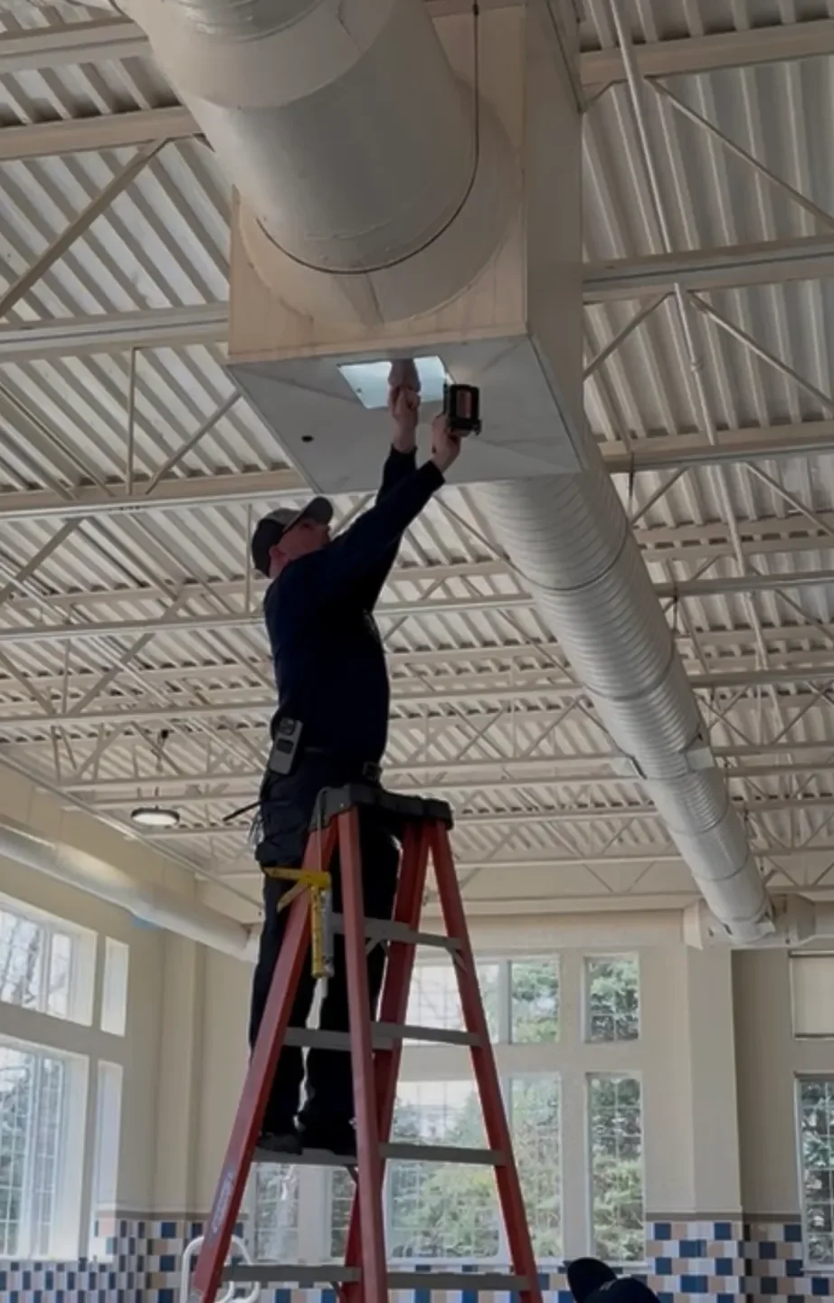 Technician cleaning commercial supply ductwork from a ladder
